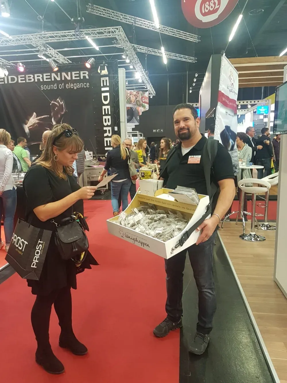 Markus Steinberger mit dem Königshappen-Bauchladen auf der Fachmesse GAST in Salzburg 2018 — reicht einer Besucherin ein Königshappen-Päckchen