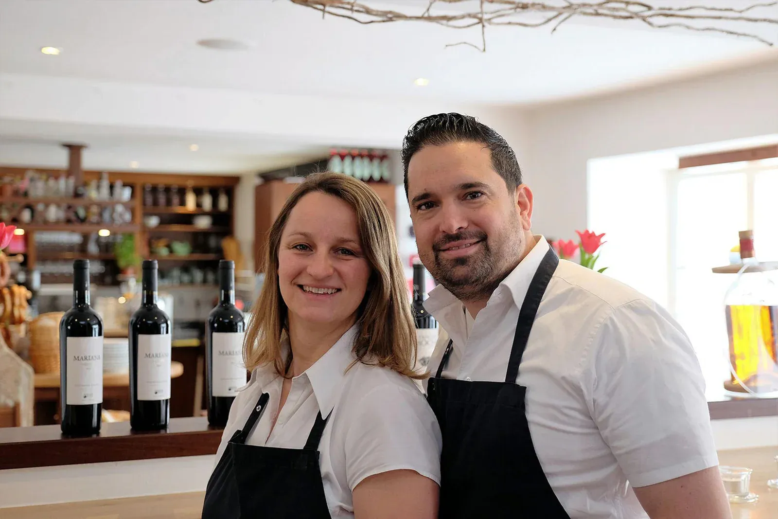 Markus und Ilonka Steinberger im Familienrestaurant Steinbergers Marktblick in Glonn — Hände am Tresen, weiße Hemden, schwarze Schürzen, Weinflaschen im Hintergrund